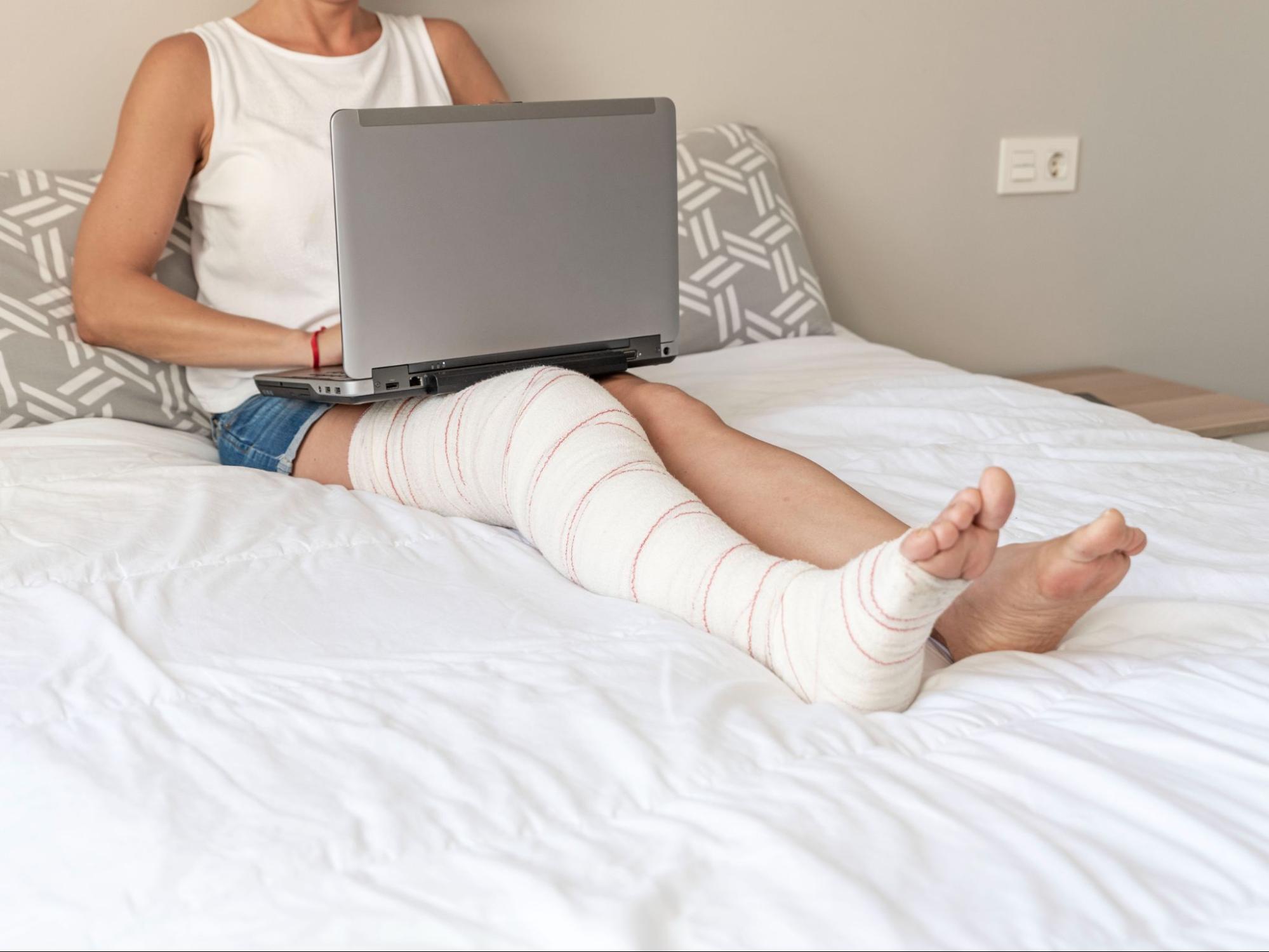Woman with leg cast working on laptop from bed